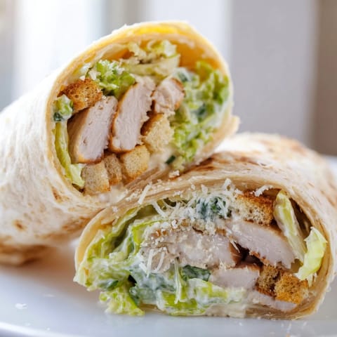 A close-up view shows a halved Caesar Chicken Salad Wrap on a plate, with creamy Caesar dressing dripping from the fresh ingredients and a side of mixed greens.